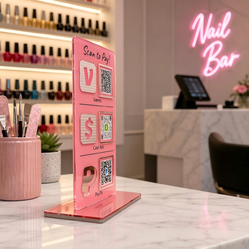 Puede incluir: Un letrero de acr&iacute;lico rosa con el texto "Scan to Pay!" y opciones de pago: Venmo, Cash App y PayPal, cada uno con un c&oacute;digo QR. El letrero est&aacute; sobre una superficie de m&aacute;rmol, con botellas de esmalte de u&ntilde;as en el fondo. Un letrero de ne&oacute;n rosa dice "Nail Bar".