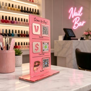 Puede incluir: Un expositor de pago de acr&iacute;lico rosa con el texto "Scan to Pay!" y c&oacute;digos QR para Venmo, Cash App y PayPal. El expositor est&aacute; sobre una encimera de m&aacute;rmol en un sal&oacute;n de manicura.