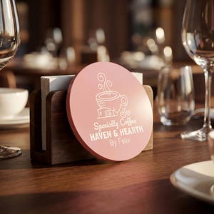 May include: A pink, circular coaster featuring a white coffee cup design and text. The coaster reads "Specialty Coffee HAVEN & HEARTH By Felix." It sits on a wooden napkin holder on a wooden table, with glasses and a white cup in the background.