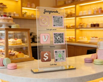 Puede incluir: Un letrero acrílico transparente con el texto "Jaaysweets_" e iconos de redes sociales para Instagram, Venmo y Cash App, cada uno con un código QR. El letrero está sobre una encimera de mármol en una panadería.