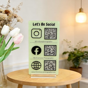 Może przedstawiać: Jasnozielony akrylowy znak mediów społecznościowych z czarnymi ikonami dla Instagrama, Facebooka i strony internetowej, każda w parze z kodem QR. Znak głosi "Let's Be Social" na górze i zawiera nazwę firmy oraz adres strony internetowej.