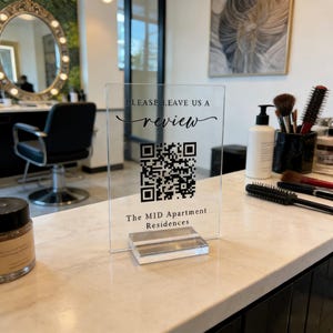May include: A clear acrylic sign with the text "PLEASE LEAVE US A review" and a QR code. Below the QR code is the text "The MID Apartment Residences". The sign is on a clear acrylic base, displayed on a marble countertop.