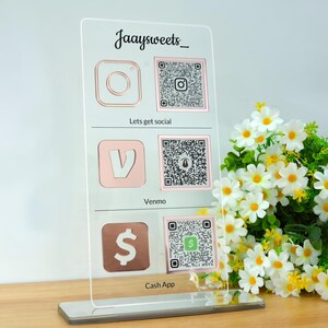 Op de afbeelding: Een helder acryl social media bord met ros&eacute;gouden iconen voor Instagram, Venmo en Cash App, elk met een QR-code. Het bord heeft de tekst "Jaaysweets_" bovenaan en de zinnen "Lets get social" en "Cash App" eronder.