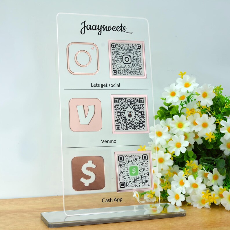 Op de afbeelding: Een helder acryl social media bord met ros&eacute;gouden iconen voor Instagram, Venmo en Cash App, elk met een QR-code. Het bord heeft de tekst "Jaaysweets_" bovenaan en de zinnen "Lets get social" en "Cash App" eronder.