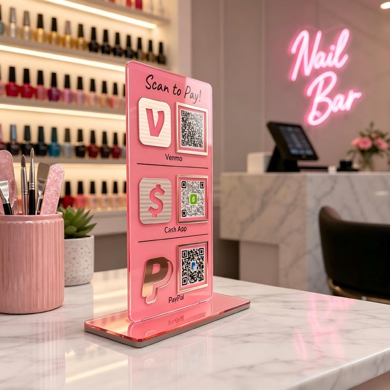 Puede incluir: Un letrero acr&iacute;lico rosa con opciones de pago para un sal&oacute;n de u&ntilde;as. El letrero muestra c&oacute;digos QR para Venmo, Cash App y PayPal, con el texto "Scan to Pay!" en la parte superior. El fondo incluye botellas de esmalte de u&ntilde;as y un letrero de ne&oacute;n "Nail Bar".
