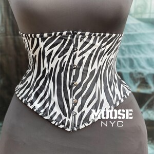 May include: A black and white zebra print corset with a pointed bottom edge. The corset has a row of silver-colored buttons down the front and is displayed on a mannequin. The text "MOOSE NYC" is printed on the bottom right.