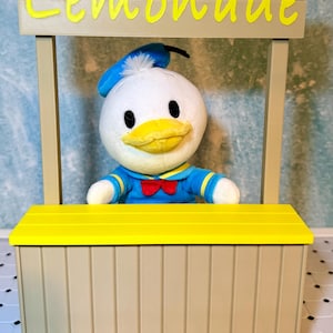 May include: A plush Donald Duck toy stands behind a yellow and tan lemonade stand. The sign above the stand reads "Lemonade" in yellow letters. Donald Duck is wearing a blue sailor suit and a red bow tie.
