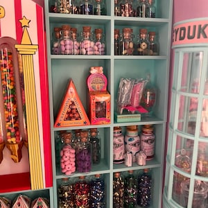 Puede incluir: Una estantería de exhibición de caramelos azul claro llena de frascos de vidrio con caramelos y golosinas de colores. La estantería incluye cajas de dulces y un dispensador de caramelos. El texto "Exploding Bonbons" es visible en una caja triangular.