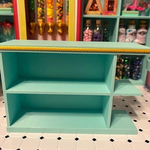Puede incluir: Una estantería de exhibición de color verde azulado claro con dos estantes y un pequeño estante lateral. El borde superior está adornado con un diseño de cuerda dorada. La estantería forma parte de una exhibición de tienda de golosinas, con caramelos y frascos coloridos en el fondo.