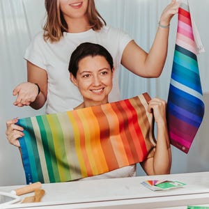 May include: Two women hold color swatches in front of a mirror. One woman holds a scarf with vertical stripes in a rainbow of colors. The other woman holds a color palette with pinks, blues, and greens. The scene is brightly lit.