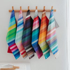 May include: A collection of seven colorful, striped towels hanging on a wooden rack. Each towel features a gradient of colors, including blue, green, pink, and orange. The towels are neatly arranged, creating a vibrant display against a white wall.