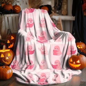 May include: A pink and white Halloween-themed throw blanket featuring a repeating pattern of cartoon ghosts reading books. The blanket is draped over a chair, with carved pumpkins in the background.