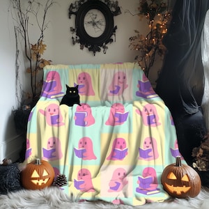 May include: A colorful blanket featuring a repeating pattern of pink ghosts reading books. The blanket is draped over a couch, with a black cat sitting on it. Two carved pumpkins and decorative branches complete the Halloween theme.