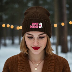 Pode incluir: Um gorro castanho com o texto "SMUT READER" e um desenho de um fantasma. O gorro é feito de um material de malha macia. O fundo está desfocado com luzes bokeh.