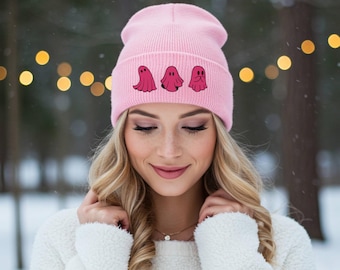Puede incluir: Un gorro rosa claro con tres ilustraciones de fantasmas rosas. El gorro está hecho de un material de punto. El fondo es una escena invernal borrosa con luces bokeh.