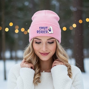 May include: A pale pink beanie hat featuring the words "SMUT READER" and a cartoon ghost with a book. The beanie is made of a soft material, ideal for colder weather. A fashionable accessory.