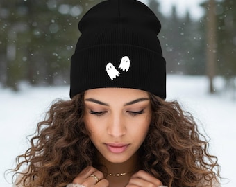 Puede incluir: Un gorro negro con un diseño de fantasmas blancos. El gorro está hecho de un tejido de punto acanalado suave. El gorro lo lleva una persona con pelo largo y rizado de color marrón. El fondo es un bosque nevado.