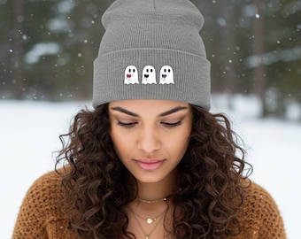 Puede incluir: Un gorro gris con tres fantasmas de dibujos animados con diferentes expresiones. Uno saca la lengua, otro tiene un corazón en el ojo y el último un corazón morado. El gorro está hecho de punto acanalado.