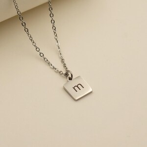 May include: Silver-toned necklace with a delicate chain and a square pendant. The pendant features the letter "m" in a minimalist font. The necklace is displayed on a neutral background, highlighting its simple elegance.