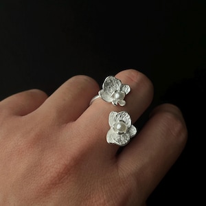 Op de afbeelding: Twee zilveren ringen, elk met een bloemdesign en een centrale parel. De ringen worden op een hand gedragen tegen een zwarte achtergrond. De bloemblaadjes hebben een getextureerd uiterlijk.