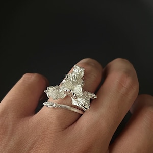 Puede incluir: Un anillo plateado con un diseño floral detallado. El anillo se envuelve alrededor del dedo con una flor calada texturizada y una banda delgada. El anillo se lleva en un dedo sobre un fondo oscuro.