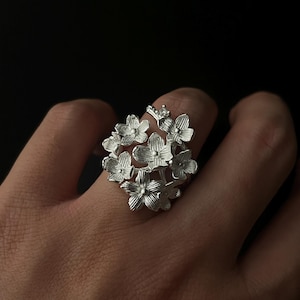 以下が含まれることがあります： 複数の花が特徴の、銀色のリング。リングは指に装着されており、暗い背景に映えています。花は細部までこだわって作られており、花のクラスターを形成しています。