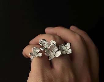 May include: Silver-toned floral rings. The rings feature detailed, openwork flower designs, with multiple rings worn on the fingers. The flowers have textured petals and are arranged to create a natural, organic look. The background is a solid black.
