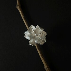 May include: A silver flower ring with a textured finish and a small, pearl-like centre. The ring is displayed on a thin, brown branch against a black backdrop. The flower has five petals.