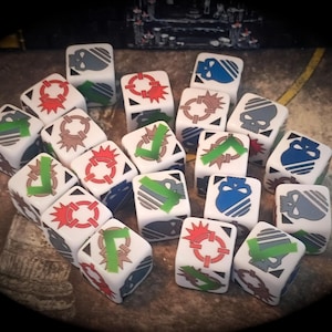 Op de afbeelding: Een verzameling witte dobbelstenen met diverse gekleurde symbolen, waaronder schedels, doelen en pijlen. De dobbelstenen zijn gerangschikt op een bruin oppervlak, wat wijst op een tabletop game. De symbolen zijn in blauw, groen, rood en bruin.