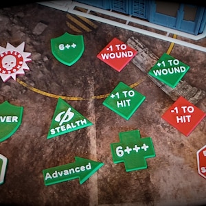 May include: A collection of colorful game tokens with various symbols and text. Green shield-shaped tokens read "COVER" and "6++." Red diamond-shaped tokens say "-1 TO WOUND" and "-1 TO HIT." Other tokens include "Sticky OBJ" and "Advanced."