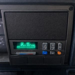 May include: Close-up of a vehicle's dashboard with a black textured panel. A digital display shows "A/C REC LOW" and temperature. Below are buttons for air conditioning, blend, and fan speed. The dashboard is dark gray.