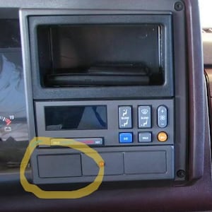 May include: A vehicle dashboard with a dark grey console. It has climate control buttons, a digital display, and a radio slot. Below the display are two rectangular storage compartments. The temperature gauge shows a range from cold to hot.