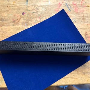 Puede incluir: Un objeto rectangular negro con una superficie texturizada, posiblemente una herramienta o un componente. Se encuentra sobre una l&aacute;mina de fieltro azul, colocada sobre una superficie de madera.