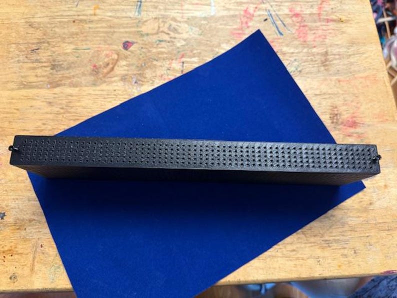 Puede incluir: Un objeto rectangular negro con una superficie texturizada, posiblemente una herramienta o un componente. Se encuentra sobre una l&aacute;mina de fieltro azul, colocada sobre una superficie de madera.