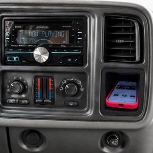 May include: A car dashboard featuring a Kenwood stereo showing "NOW PLAYING". A smartphone in a red case is in a compartment below the air vent. Climate control knobs are also visible.