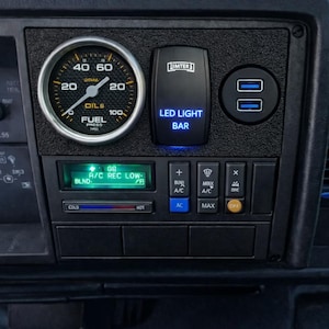 May include: Close-up of a vehicle dashboard with gauges and controls. Features a fuel pressure gauge, a digital display, and a switch labeled "LED LIGHT BAR." Includes USB ports and climate control buttons. The dashboard is black.