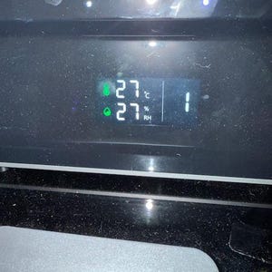 Puede incluir: Una pantalla digital que muestra la temperatura a 27 grados Celsius y la humedad al 27%. La pantalla tambi&eacute;n muestra el n&uacute;mero 1. La pantalla est&aacute; sobre una superficie negra.