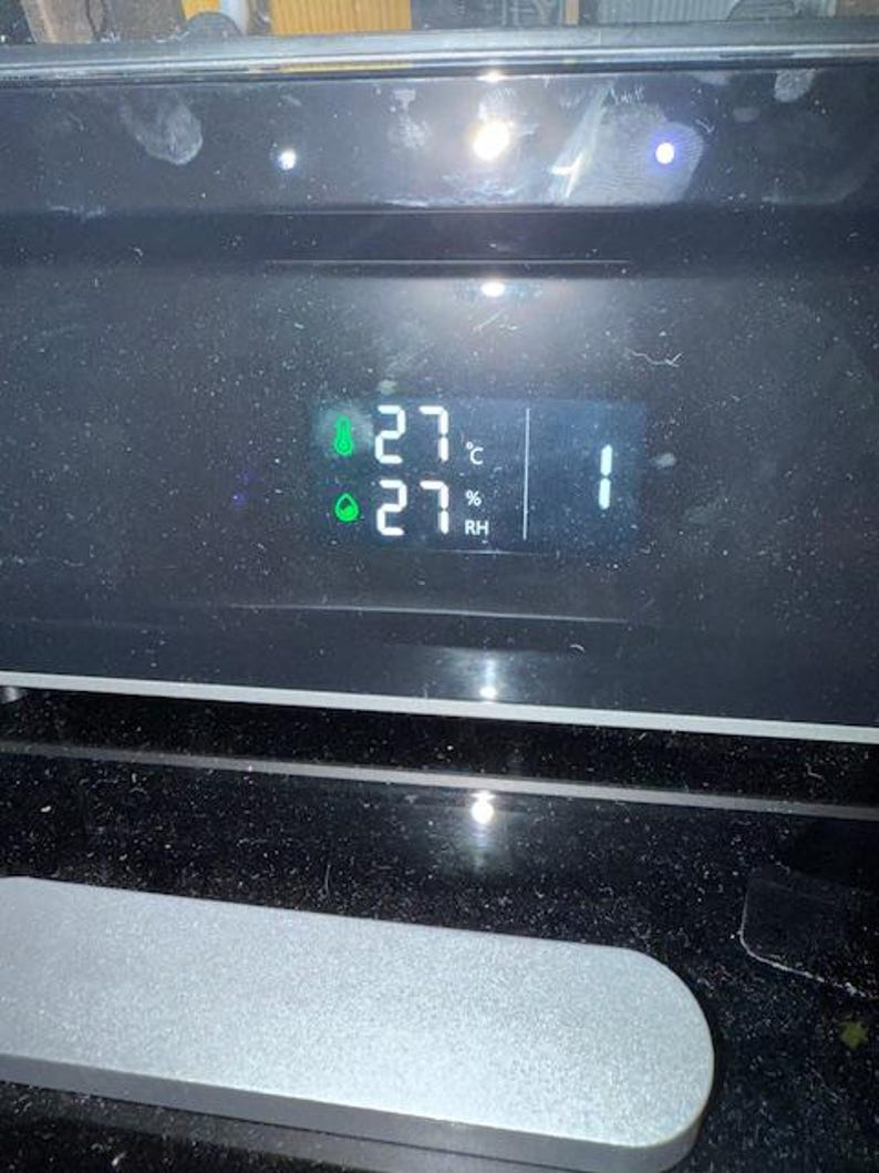 Puede incluir: Una pantalla digital que muestra la temperatura a 27 grados Celsius y la humedad al 27%. La pantalla tambi&eacute;n muestra el n&uacute;mero 1. La pantalla est&aacute; sobre una superficie negra.