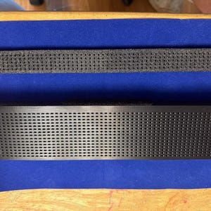 Puede incluir: Objeto rectangular negro con superficie perforada, sobre tela azul. Encima, un objeto negro perforado similar m&aacute;s peque&ntilde;o. Los objetos est&aacute;n sobre una superficie de madera.