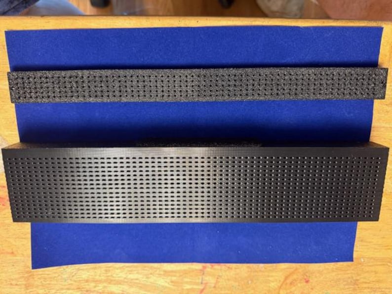 Puede incluir: Objeto rectangular negro con superficie perforada, sobre tela azul. Encima, un objeto negro perforado similar m&aacute;s peque&ntilde;o. Los objetos est&aacute;n sobre una superficie de madera.