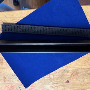Puede incluir: Se muestran dos objetos rectangulares negros. Uno tiene una superficie texturizada y se encuentra encima de un objeto rectangular hueco. Ambos est&aacute;n sobre una superficie triangular azul, sobre un fondo de madera.
