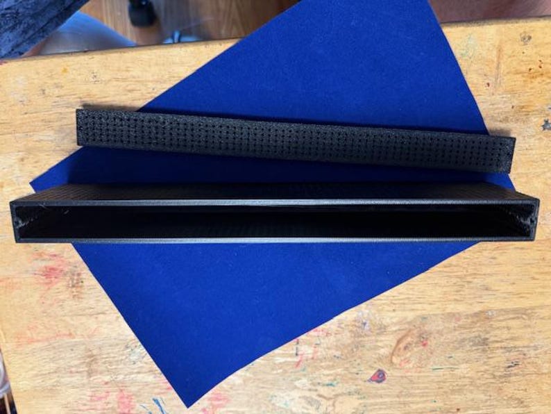 Puede incluir: Se muestran dos objetos rectangulares negros. Uno tiene una superficie texturizada y se encuentra encima de un objeto rectangular hueco. Ambos est&aacute;n sobre una superficie triangular azul, sobre un fondo de madera.