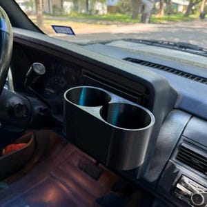 May include: A black dual cup holder accessory mounted onto the dashboard of a vehicle. The unit features two circular openings for holding beverages and is positioned directly in front of the steering column and instrument cluster area.