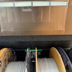 Puede incluir: Un contenedor de almacenamiento de filamento para impresora 3D con m&uacute;ltiples bobinas de filamento. El contenedor est&aacute; abierto, revelando varias bobinas de filamento en varios colores, incluyendo gris, negro y azul. El contenedor tiene una parte superior perforada.