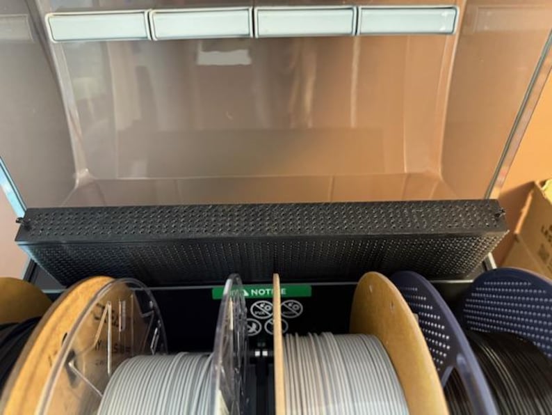 Puede incluir: Un contenedor de almacenamiento de filamento para impresora 3D con m&uacute;ltiples bobinas de filamento. El contenedor est&aacute; abierto, revelando varias bobinas de filamento en varios colores, incluyendo gris, negro y azul. El contenedor tiene una parte superior perforada.