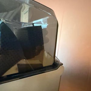 Puede incluir: Primer plano de un electrodom&eacute;stico transparente cubierto de pl&aacute;stico con un componente rectangular negro en el interior. El electrodom&eacute;stico tiene una base de color claro y una parte superior oscura y curvada. El fondo es una pared marr&oacute;n claro.