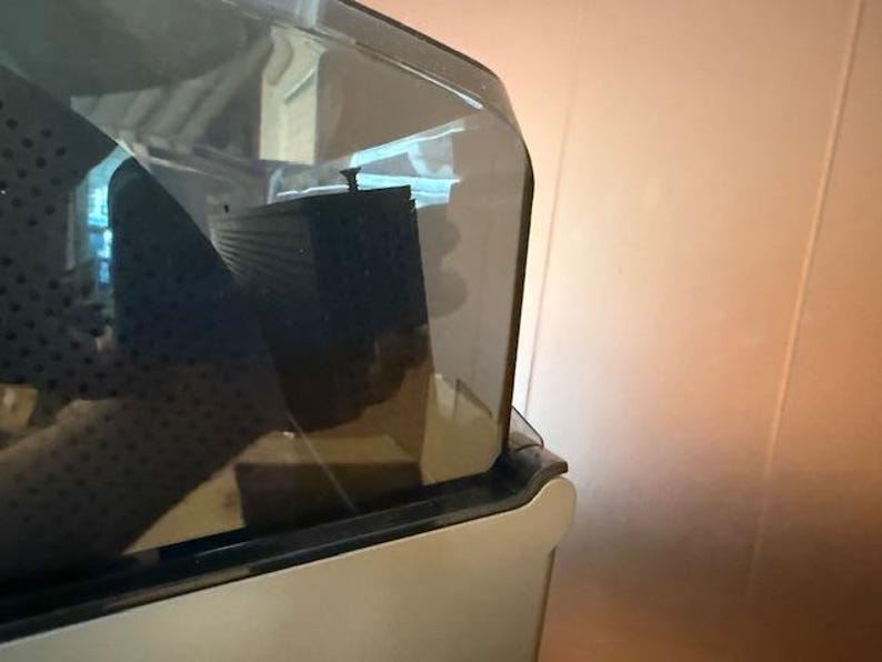 Puede incluir: Primer plano de un electrodom&eacute;stico transparente cubierto de pl&aacute;stico con un componente rectangular negro en el interior. El electrodom&eacute;stico tiene una base de color claro y una parte superior oscura y curvada. El fondo es una pared marr&oacute;n claro.