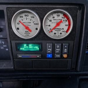 May include: Car dashboard featuring two Pro-Comp gauges displaying oil and nitrous pressure. The gauges have white faces, red needles, and black text. Below are A/C controls and a digital display.
