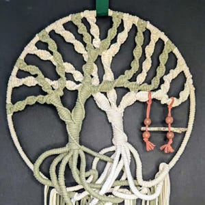 May include: Macrame wall hanging featuring a tree of life design. The artwork uses off-white and olive green cords to create the tree and its roots. A small swing hangs from the branches. The piece is finished with a green ribbon for hanging.