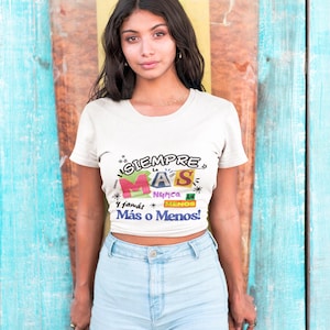 Op de afbeelding: Een vrouw draagt een witte crop top met de Spaanse tekst "Siempre Más, Nunca Menos! Más o Menos!" in kleurrijke letters. Ze draagt een lichtblauwe spijkerbroek. De achtergrond is blauw en geel.